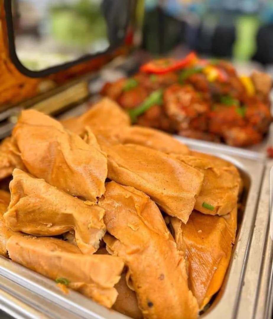 Leaf-wrapped moi moi in a chafing dish with peppered chicken behind it