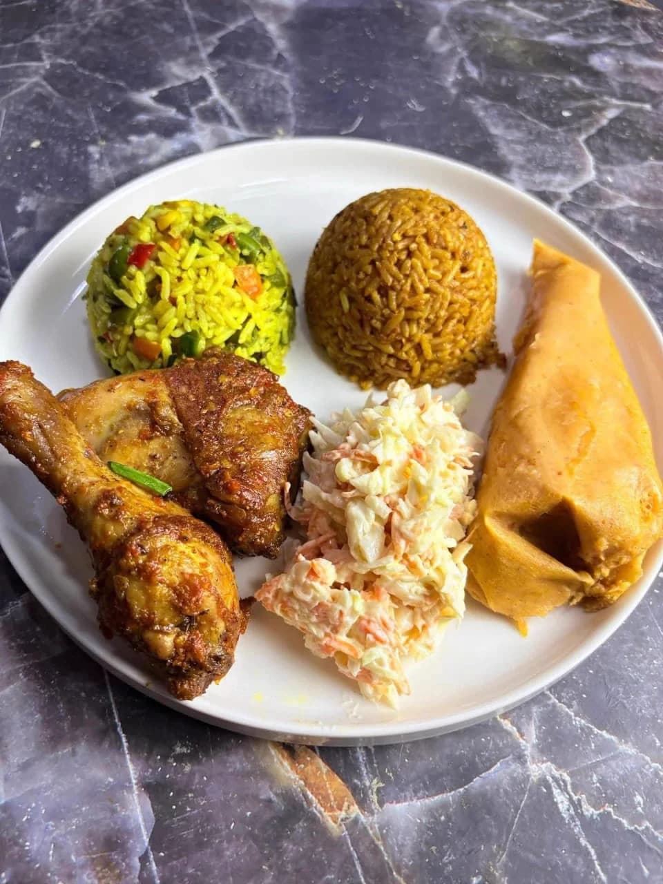A plated meal of peppered chicken, jollof rice, fried rice, coleslaw and swallow